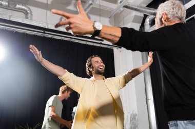 excited bearded man with outstretched hands rehearsing near grey haired art director in theater 