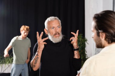 tattooed and bearded art director gesturing while talking to blurred actor in theater. Translation of tattoo: 