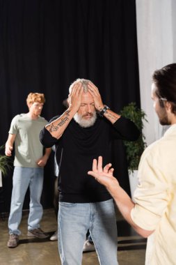 frustrated art director with closed eyes touching head near blurred gesturing actor. Translation of tattoo: peace