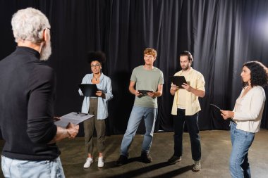 young interracial actors holding clipboards near grey haired screenwriter on blurred foreground clipart