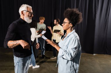 african american actress with clipboard looking at screenwriter near actors in acting school. Translation of tattoo: om, shanti, peace