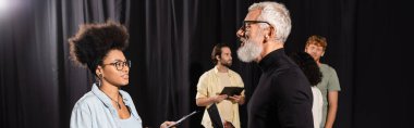bearded art director talking to african american actress near students on background, banner