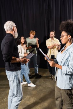 multiethnic actors holding clipboard with scenarios near grey haired screenwriter in theater clipart