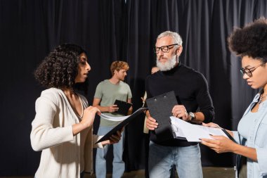 bearded acting skills teacher looking at interracial students holding clipboards with screenplays clipart