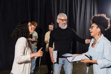 cheerful african american woman holding screenplay and laughing near bearded producer and multiethnic actors in theater clipart