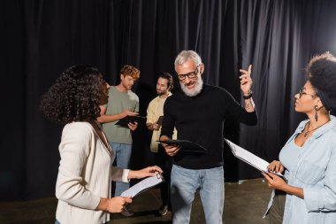 bearded art director pointing with finger while reading screenplay near multiethnic actresses during acting skills lesson clipart