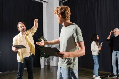 redhead and bearded actors holding clipboards with screenplays while rehearsing in theater 