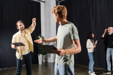 redhead and bearded actors holding clipboards with screenplays while rehearsing in theater  clipart