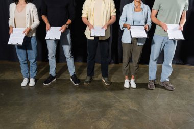 partial view of multiethnic actors holding clipboards with scenarios while standing on stage in theater clipart