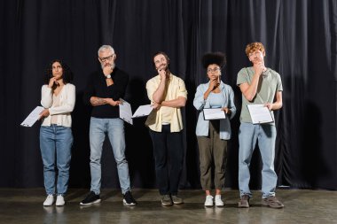 full length of thoughtful multicultural actors with bearded grey haired art director holding scenarios while rehearsing on stage in theater clipart