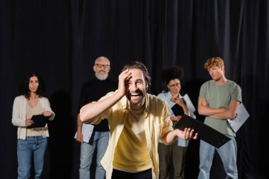 laughing man touching face and looking at camera near blurred multiethnic actors and producer in theater school