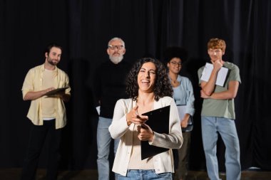 excited multiracial actress laughing and pointing with finger near multiethnic students teacher on blurred background