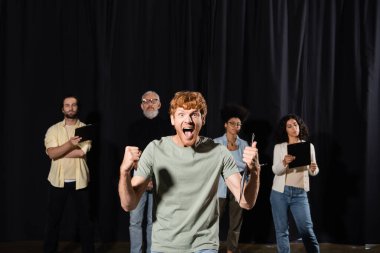 emotional redhead man showing thumb up and screaming near multiethnic students and teacher of acting skills on background clipart