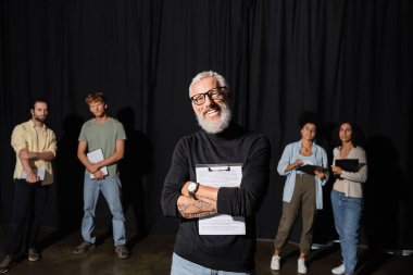 happy bearded art director holding screenplay and looking at camera near young interracial students. Translation of tattoo: om, shanti, peace