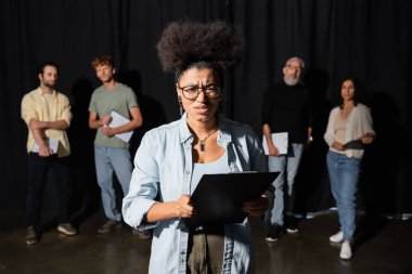 frowning african american woman holding clipboard and looking at camera near multicultural actors and producer on blurred background clipart