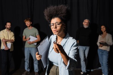 african american woman holding screenplay and gesturing during rehearsal near blurred actors and acting skills teacher