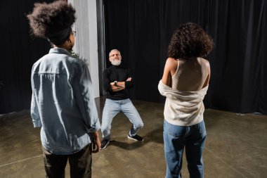 bearded teacher posing with crossed arms near multiethnic actresses in acting school. Translation of tattoo: om, shanti, peace 