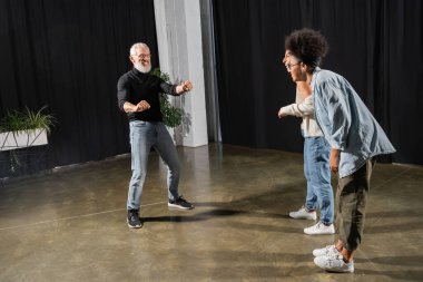 interracial actresses laughing near bearded art director posing and gesturing in theater. Translation of tattoo: om, shanti, peace 