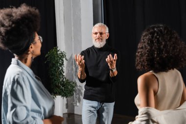 bearded acting skills teacher gesturing near blurred multiethnic actresses during lesson in theater. Translation of tattoo: om, shanti, peace