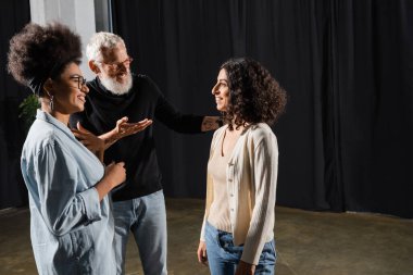 smiling art director pointing with hand and talking to multiracial woman near african american actress in theater