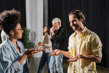 smiling brunette man talking to african american woman near interracial colleagues in acting school clipart