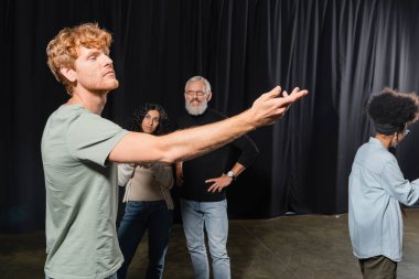 redhead man posing with outstretched hand near interracial women and smiling art director in theater clipart