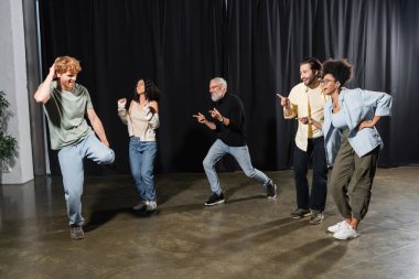 cheerful multicultural actors pointing at redhead man rehearsing on stage in theater 