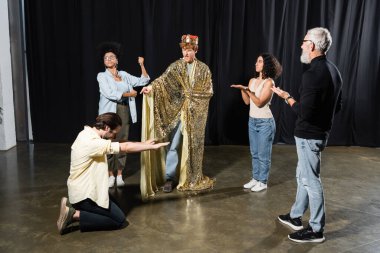 redhead man in king costume rehearsing role near multiethnic actors and art director in acting school clipart
