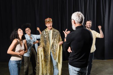 cheerful redhead man in king costume near multiethnic actors and art director on stage in theater clipart