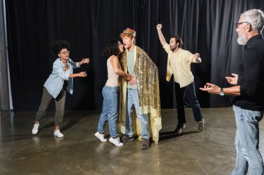 bearded producer gesturing near interracial students rehearsing scene of king assassination in theater