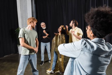 cheerful multiracial woman in queen costume smiling near interracial actors and grey haired art director. Translation of tattoo: om, shanti, peace