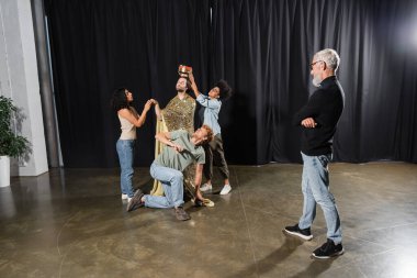 middle aged producer looking at multiethnic students rehearsing scene of king coronation on stage in theater