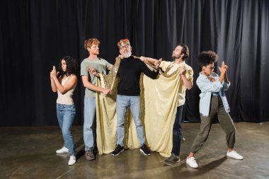 bearded art director standing in costume of king near multicultural students playing guards on stage in theater