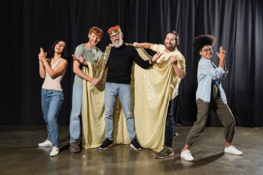 bearded art director in costume of king smiling at camera near cheerful multiethnic actors