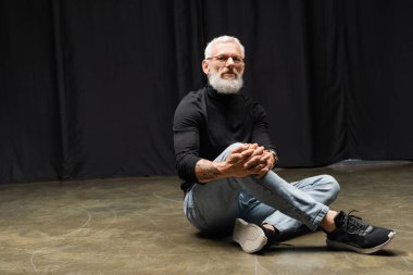 tattooed actor in eyeglasses and black turtleneck looking at camera and sitting on stage in theater. Translation of tattoo: om, shanti, peace