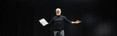 grey haired tattooed man in black turtleneck holding screenplay while rehearsing in theater, banner. Translation of tattoo: kanji, danger