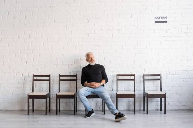 full length of mature actor in jeans looking away while sitting on chair and waiting for casting