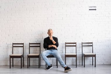 full length of pensive bearded man sitting in hall near white wall and waiting for casting. Translation of tattoo: om, shanti, peace