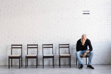 full length of pensive man in black turtleneck and jeans waiting for casting on chair near white wall