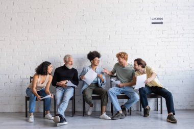 redhead man pointing with hand while talking to multicultural actors waiting for casting in hall
