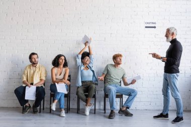 bearded art director pointing at happy african american actress holding screenplay in raised hands while sitting near interracial job seekers