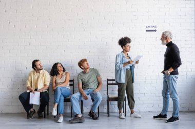 bearded art director standing with hands in pockets near african american woman reading screenplay and multiethnic actors 