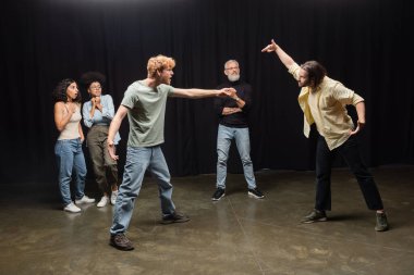full length of young actors rehearsing near astonished interracial women and art director. Translation of tattoo: om, shanti, peace  