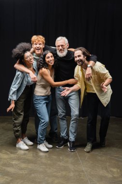 young and joyful interracial actors looking at camera while embracing bearded producer in theater