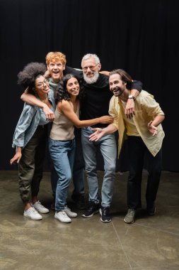 young and joyful interracial actors looking at camera while embracing bearded producer in theater clipart