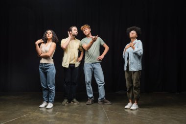 redhead man standing with hands on hips and pointing with hands near multicultural actors in acting studio