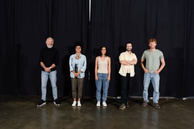 full length of happy art director with young multicultural actors standing on theater stage and looking at camera