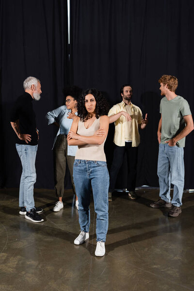 multiracial actress standing with crossed arms near interracial theater students and art director talking on background