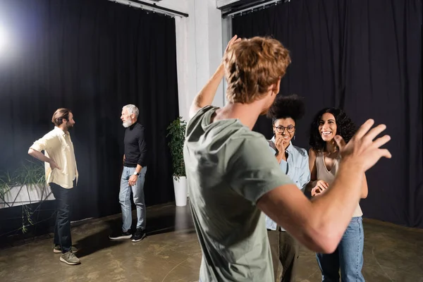 interracial actresses smiling near redhead man gesturing during rehearsing in acting skills school