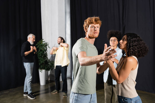 redhead man pointing with hand near interracial actresses and actors on blurred background